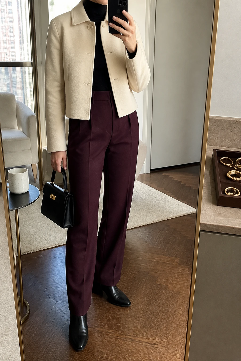 Image Prompt: Mirror selfie of a woman wearing a cream cropped tailored wool-blend jacket with a structured collar over a black fitted fine-knit turtleneck sweater, deep burgundy high-waisted straight-leg tailored trousers, black leather pointed-toe ankle boots with low block heels, a small structured black leather handbag, and delicate gold stud earrings, holding a phone in front of a mirror, cropped at the NECK so the face does not show, with a relaxed casual selfie pose, in a stylish city apartment, soft natural window light, a coffee mug on a side table and a jewelry tray on the vanity, realistic cream wool jacket texture, natural burgundy trouser folds and understated polished winter styling, latest iPhone photo quality, no additional clothing or accessories.