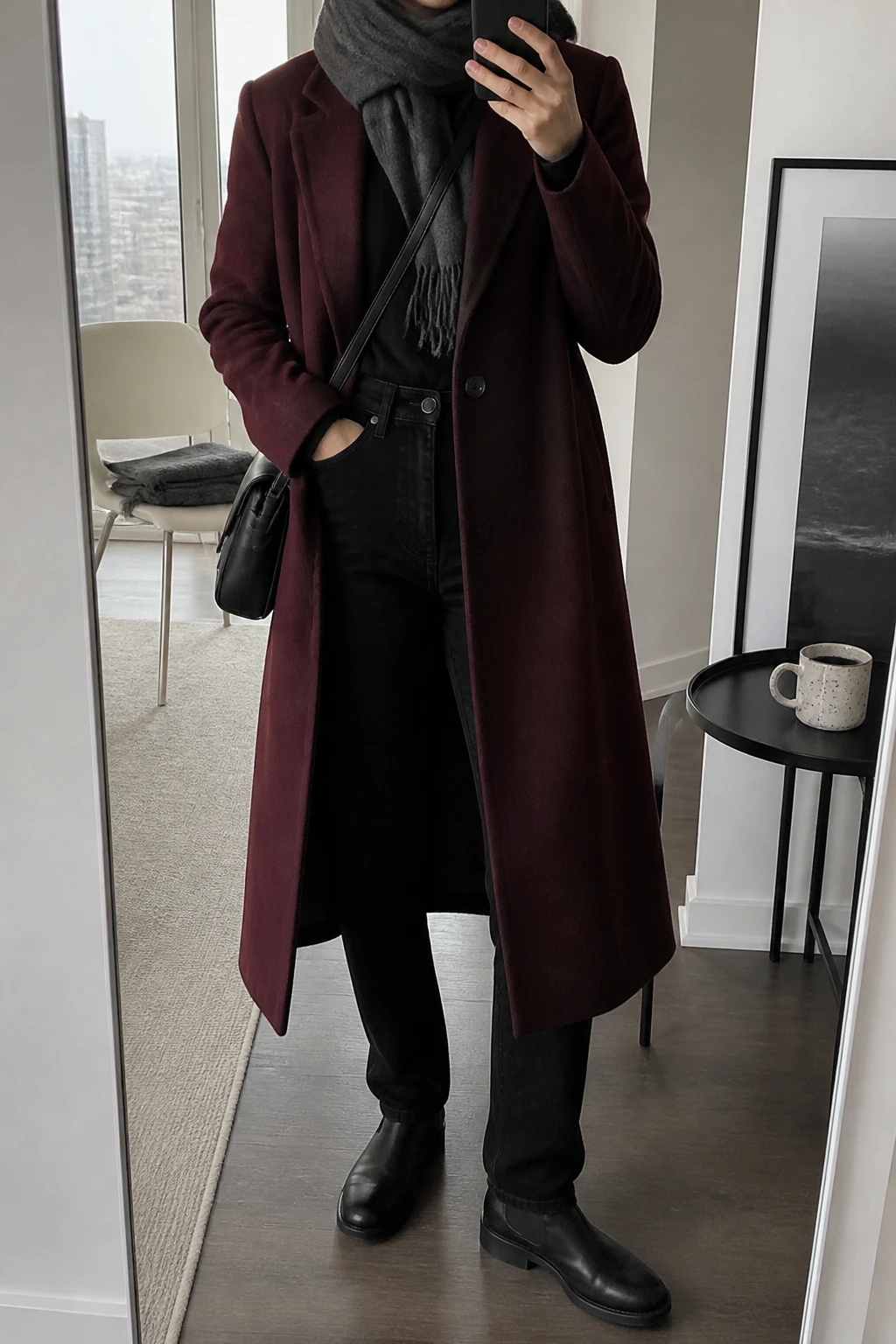 Image Prompt: Mirror selfie of a woman wearing a long burgundy single-breasted tailored wool coat over a black fitted merino wool turtleneck sweater, solid black high-waisted straight-leg denim jeans, black waterproof leather flat ankle boots with rubber soles, a structured black nylon crossbody bag, and a charcoal grey wool scarf wrapped around the neck, holding a phone in front of a mirror, cropped at the NECK so the face does not show, with a relaxed casual selfie pose, in a stylish city apartment, soft natural window light, a coffee mug on a side table and a folded blanket on a chair, realistic burgundy wool texture, natural black denim creases and soft scarf folds, latest iPhone photo quality, no additional clothing or accessories.
