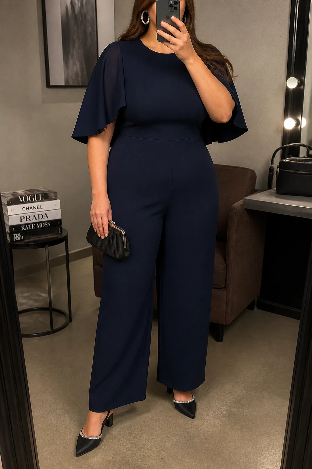 Mirror selfie of a plus size woman wearing a navy blue crepe jumpsuit with straight legs, a fitted bodice, and short cape sleeves, black crystal-trimmed slingback shoes with low block heels, a small black satin clutch, and silver crystal hoop earrings, clearly showing the exact jumpsuit shape, navy fabric, cape sleeves, footwear, and accessories listed, holding a phone in front of a mirror, cropped at the neck so her face is hidden, relaxed polished selfie pose, in a fashion studio mirror corner with warm moody lighting, a stack of fashion magazines on a side table and a cosmetic bag on the counter, realistic formal graduation styling, latest iPhone photo quality.