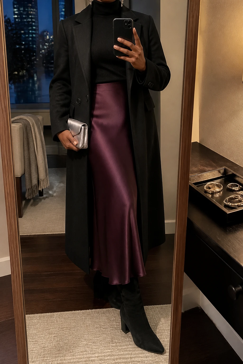 Image Prompt: Mirror selfie of a black woman wearing a black fitted fine-knit cashmere turtleneck sweater tucked into a deep plum satin bias-cut maxi skirt, black suede knee-high boots with low block heels, a long black tailored wool coat worn open, a small silver satin clutch, and delicate silver drop earrings, holding a phone in front of a full-length mirror, cropped at the NECK so the face does not show, with a relaxed casual selfie pose, in a stylish city apartment, warm moody lighting, a jewelry tray on the vanity and a folded blanket on a chair, realistic deep plum satin fabric with subtle natural reflections, soft black cashmere texture and polished evening proportions, latest iPhone photo quality, no additional clothing or accessories.