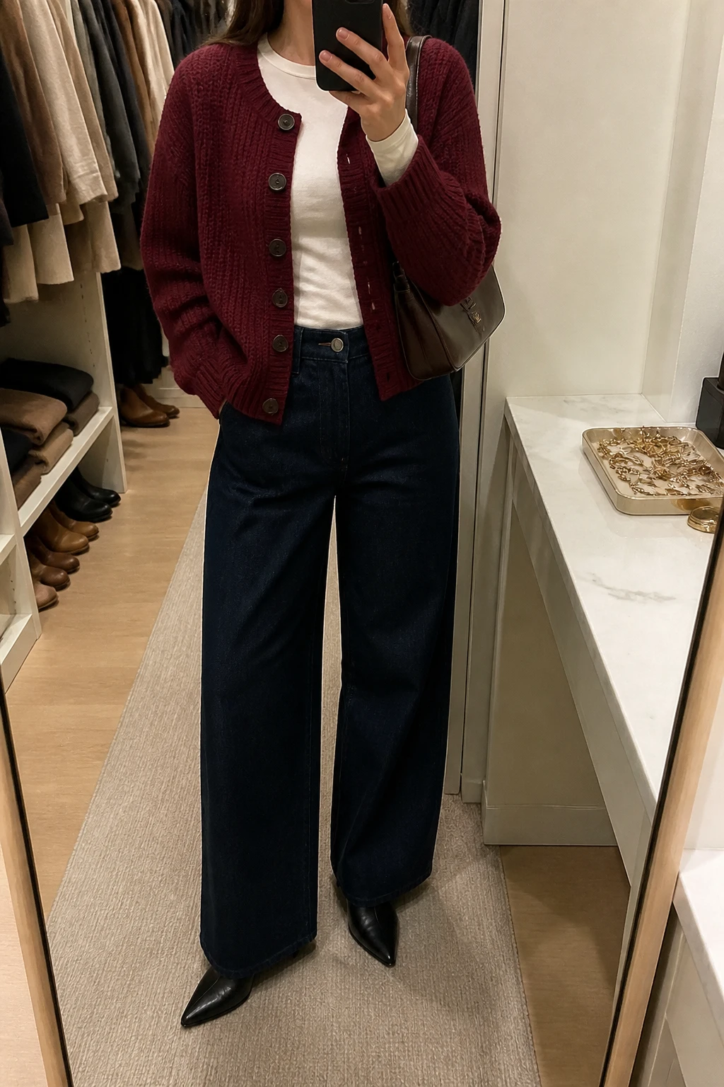 Image Prompt: Mirror selfie of a woman wearing a burgundy chunky-knit button-front cardigan over an ivory fitted cotton long-sleeve crewneck top, dark indigo high-waisted wide-leg denim jeans, black leather pointed-toe low-heeled ankle boots, and a small chocolate brown leather shoulder bag, holding a phone in front of a mirror, cropped at the NECK so the face does not show, with a relaxed casual selfie pose, in a walk-in closet mirror, warm soft indoor lighting, an open closet with clothes visible and a jewelry tray on the vanity, realistic burgundy cardigan texture, natural dark denim folds and understated autumn-to-winter styling, latest iPhone photo quality, no additional clothing or accessories.